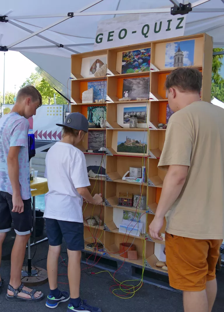 Drei Personen stehen vor einm Holzregal mit vielen Fächern. Diese enthalten Bilder, Steine und andere Gegenstände. Die Fächer lassen sich mit Kabeln verbinden und sind an eine Blaulicht-Lampe angeschlossen.