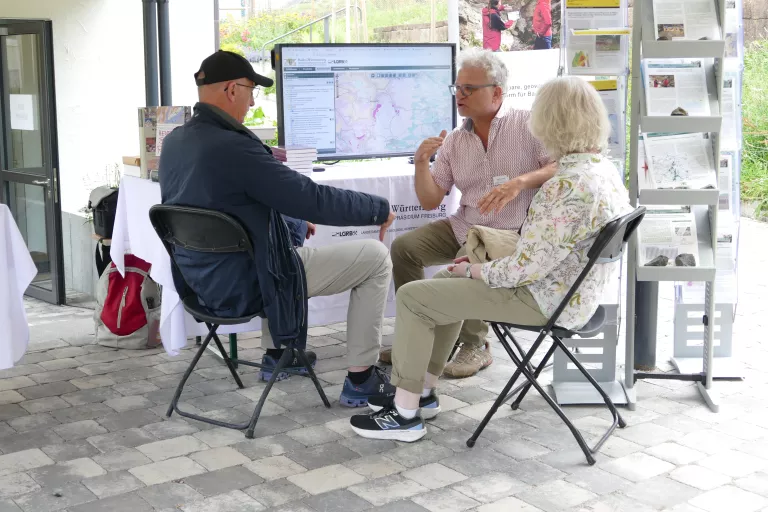 Vor einem großen Bildschirm sitzen 3 Personen und diskutieren