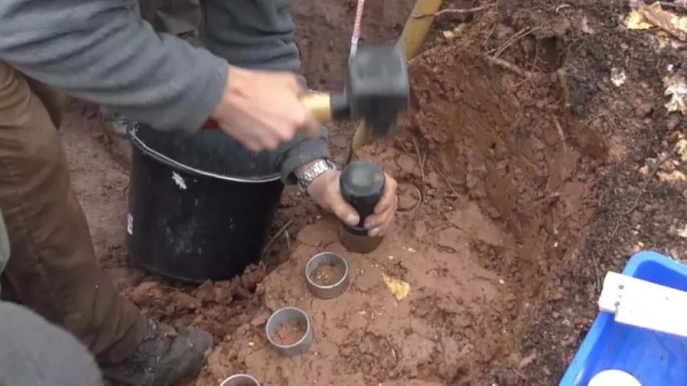 In einer in rötlichem Tonboden aufgegrabenen Profilgrube werden rundliche Metallzylinder eingeschlagen, zur Entnahme von Bodenproben.