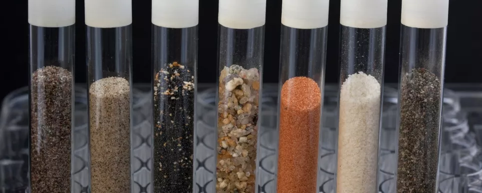 Sieben Reagenzgläser stehen in einem Reagenzglashalter aus Kunststoff. Die Reagenzgläser sind mit Proben aus Sand verschiedener Herkunft befüllt. Sand ist eine Korngröße, die petrographische Zusammensetzung kann unterschiedlich sein. Die Sandproben variieren in ihrer Farbe von grau, beige, schwarz, ocker, orange bis weiß.